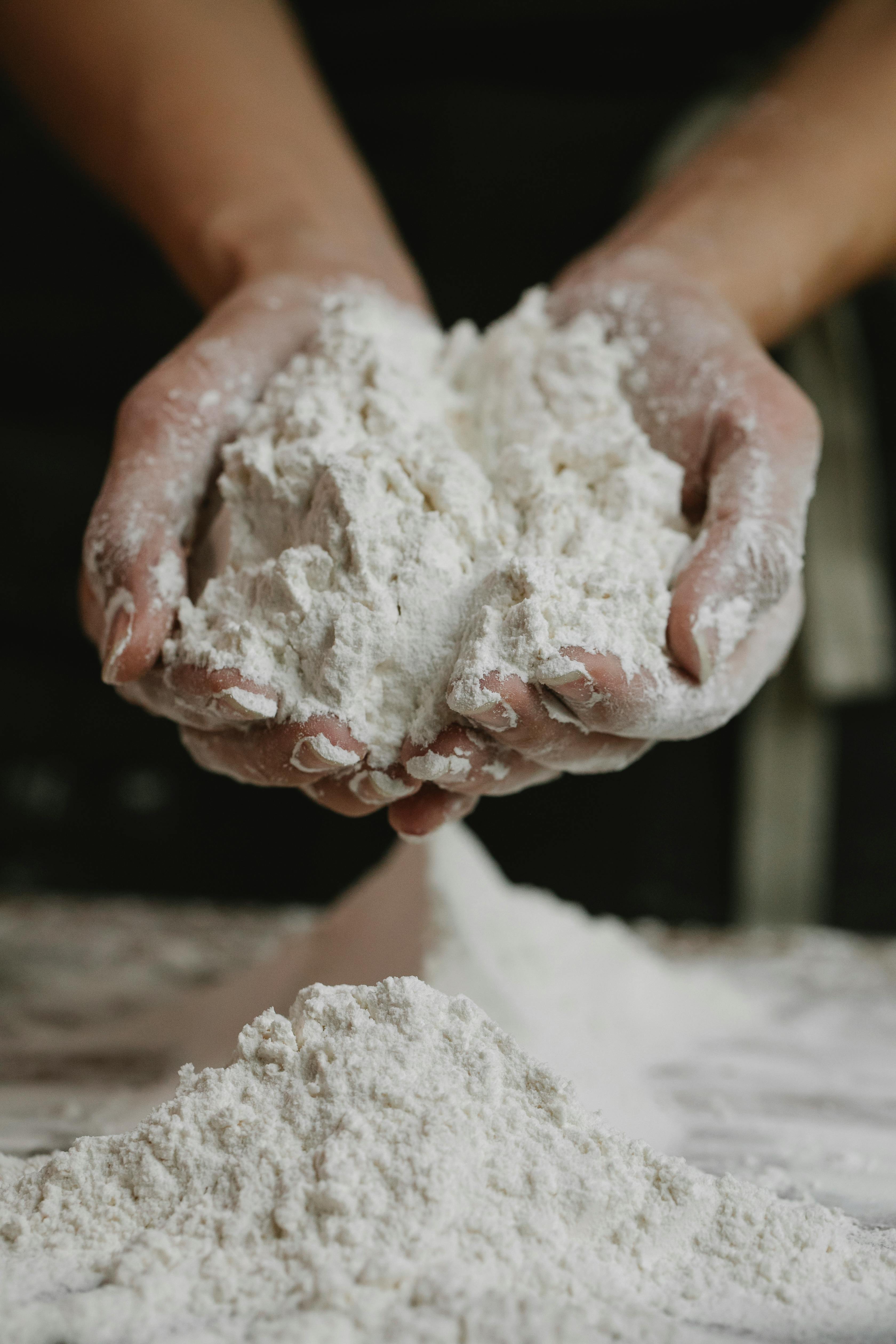Nos Produits Close-up of a person holding a mound of flour in their hands inside a kitchen.